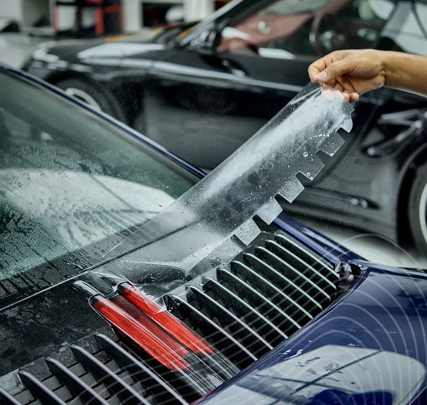 An Apex installer lifting clear paint protection film over a Porsche engine grille